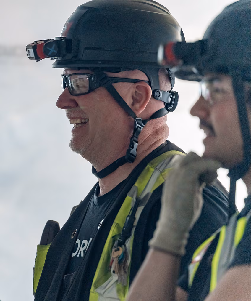 Two construction workers wearing safety helmets, glasses, and high-visibility vests, one smiling.