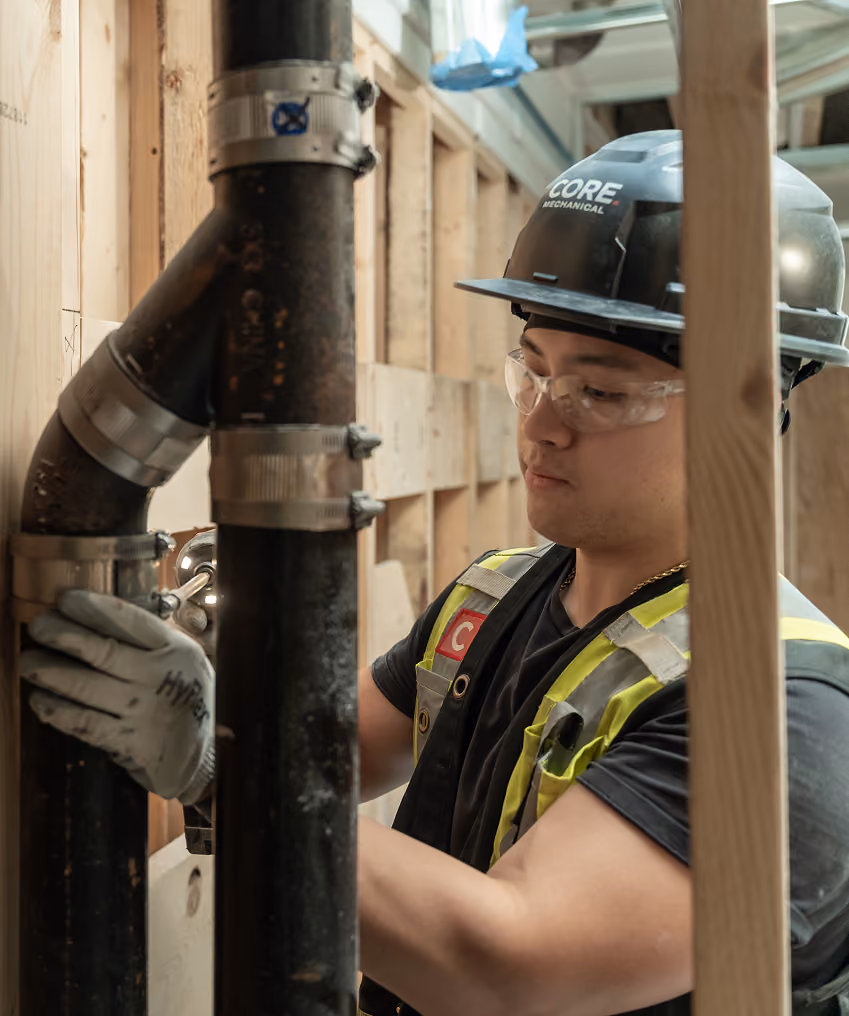 Construction worker wearing safety gear fixing pipes inside a wooden framed building.