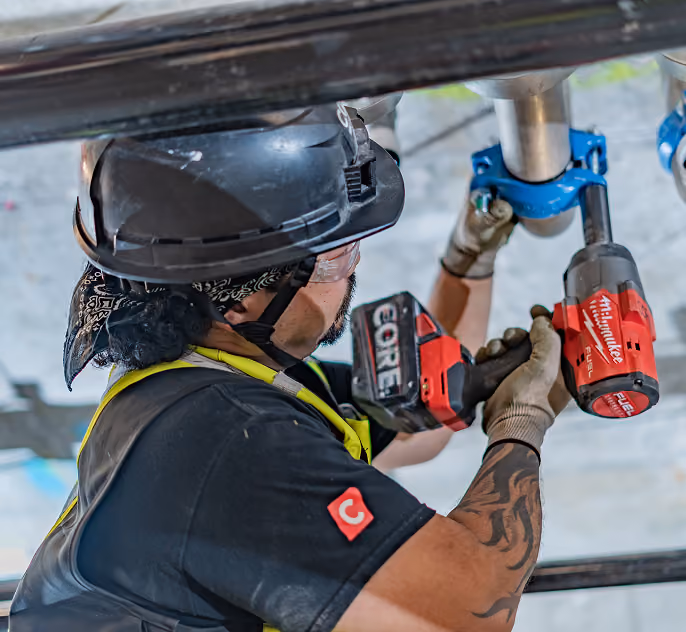 Construction worker wearing helmet and gloves using a red power tool to tighten a pipe fitting overhead.