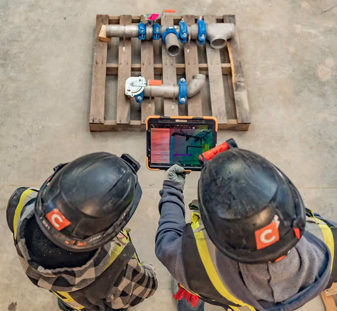 Two workers in helmets using a tablet to inspect metal pipes on a wooden pallet.