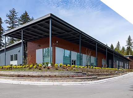 Modern single-story building with large windows, metal roof, and landscaped garden under a blue sky.