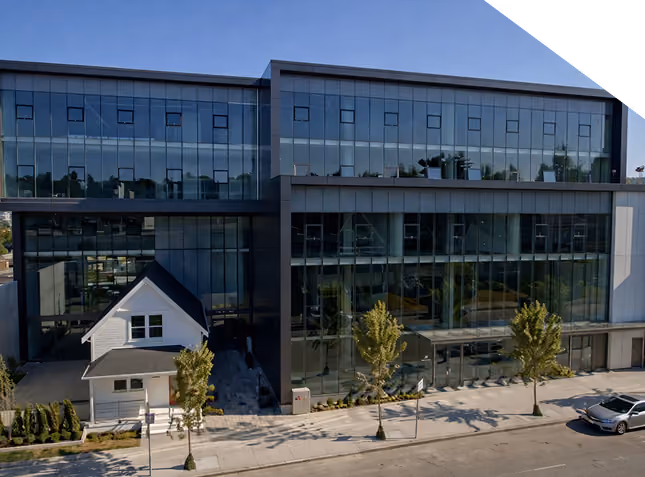 Modern glass office building with a small white house at its base and a parked car nearby.