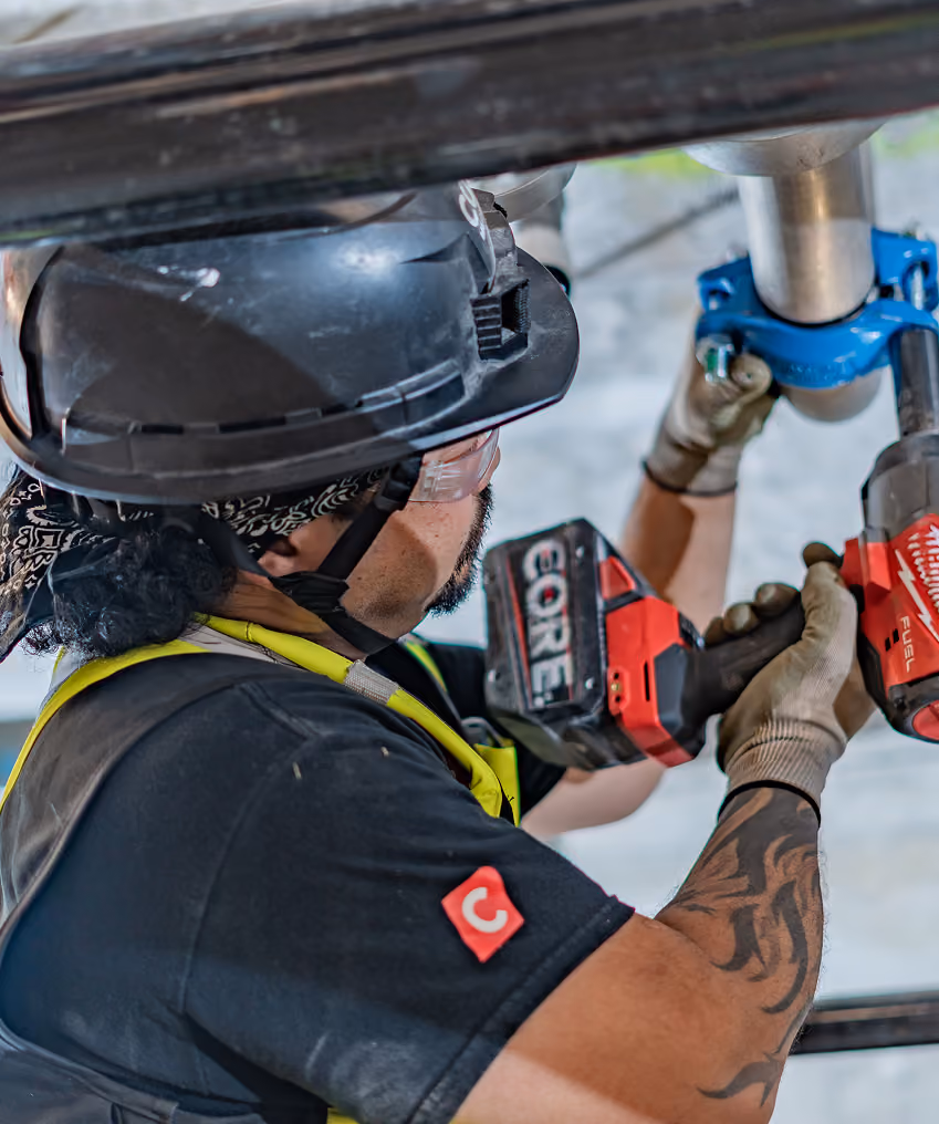 Construction worker with hard hat using a power tool on metal piping indoors.