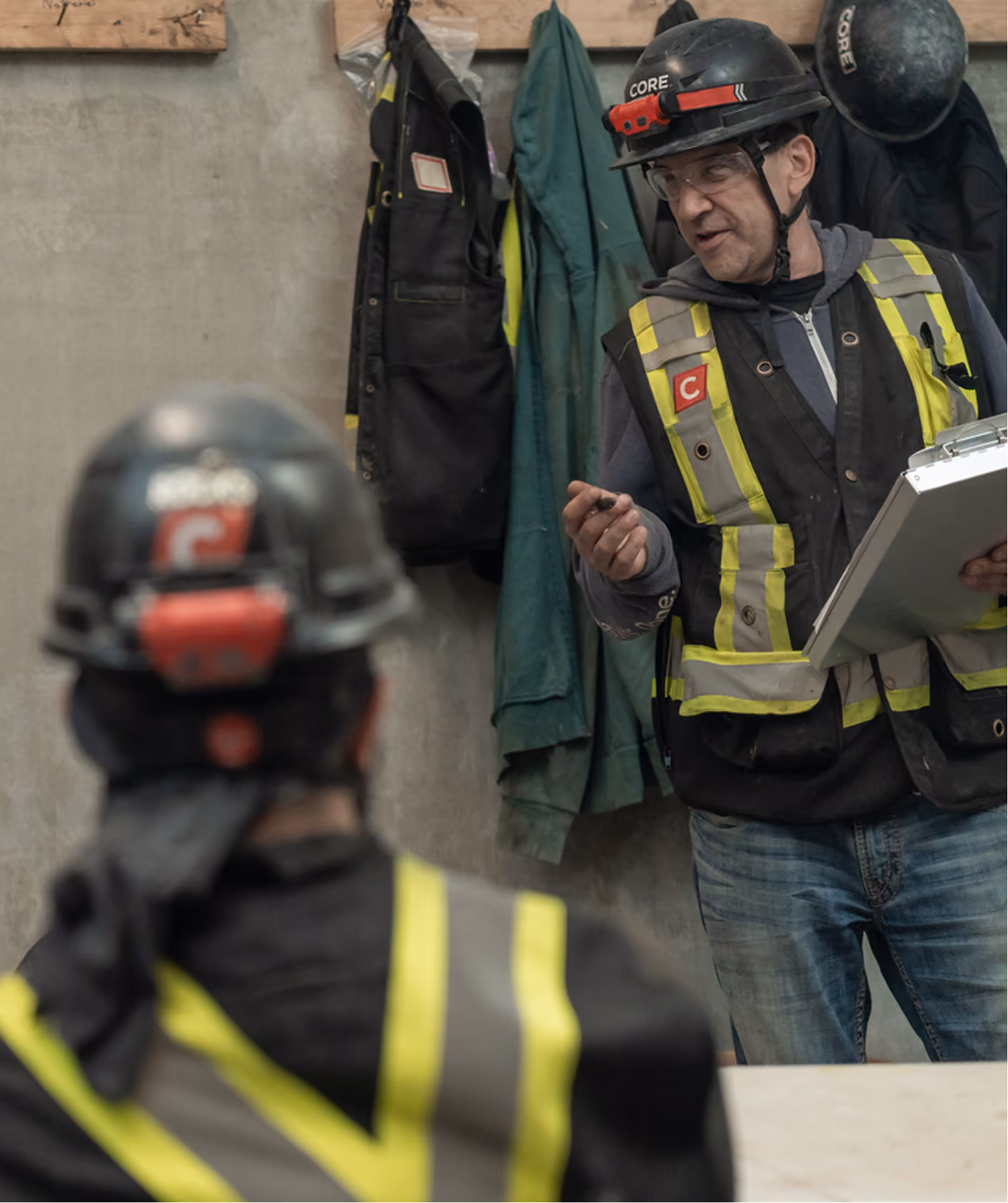 Construction workers wearing helmets and safety vests having a discussion indoors.