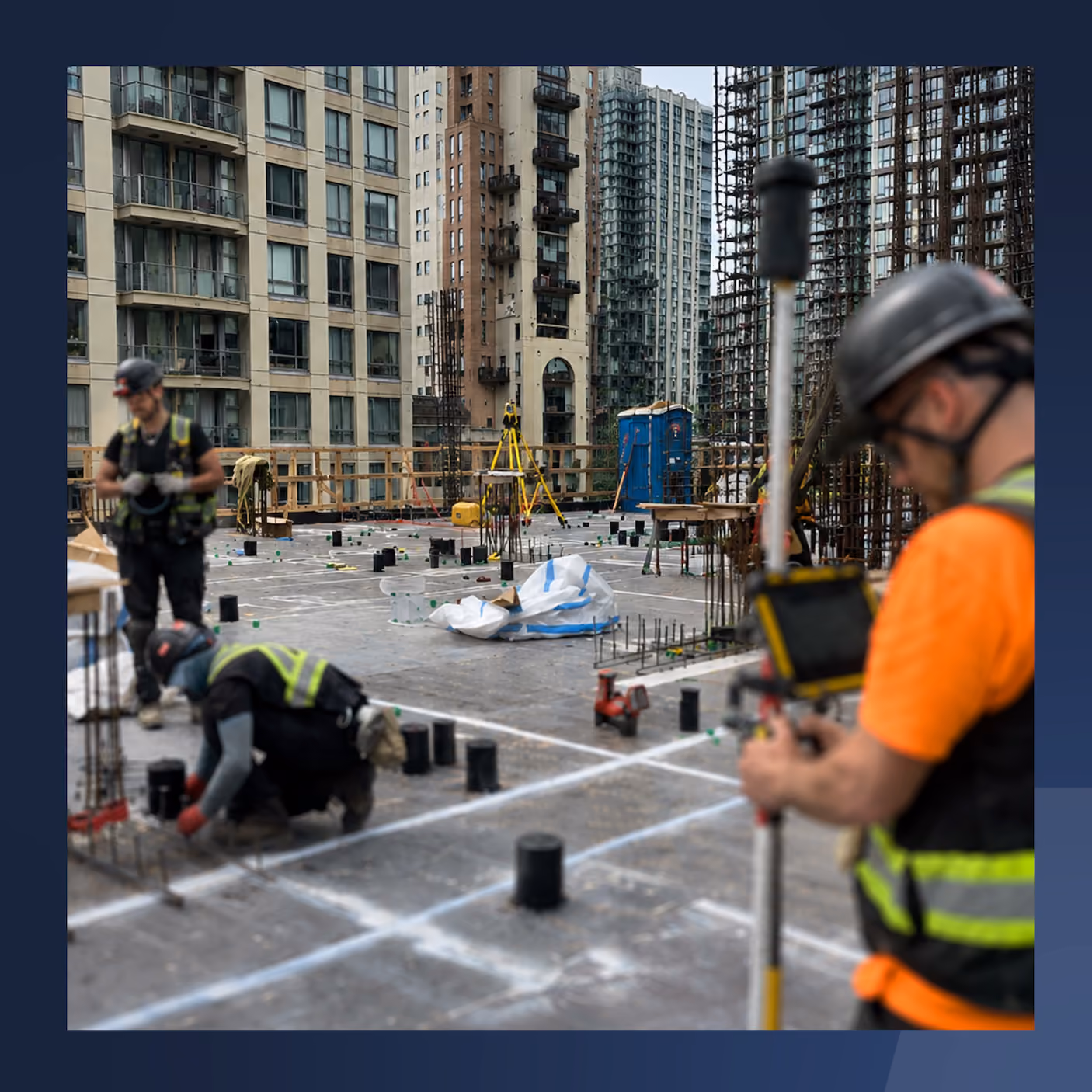 Construction workers wearing helmets and vests working on a high-rise building foundation in an urban area.