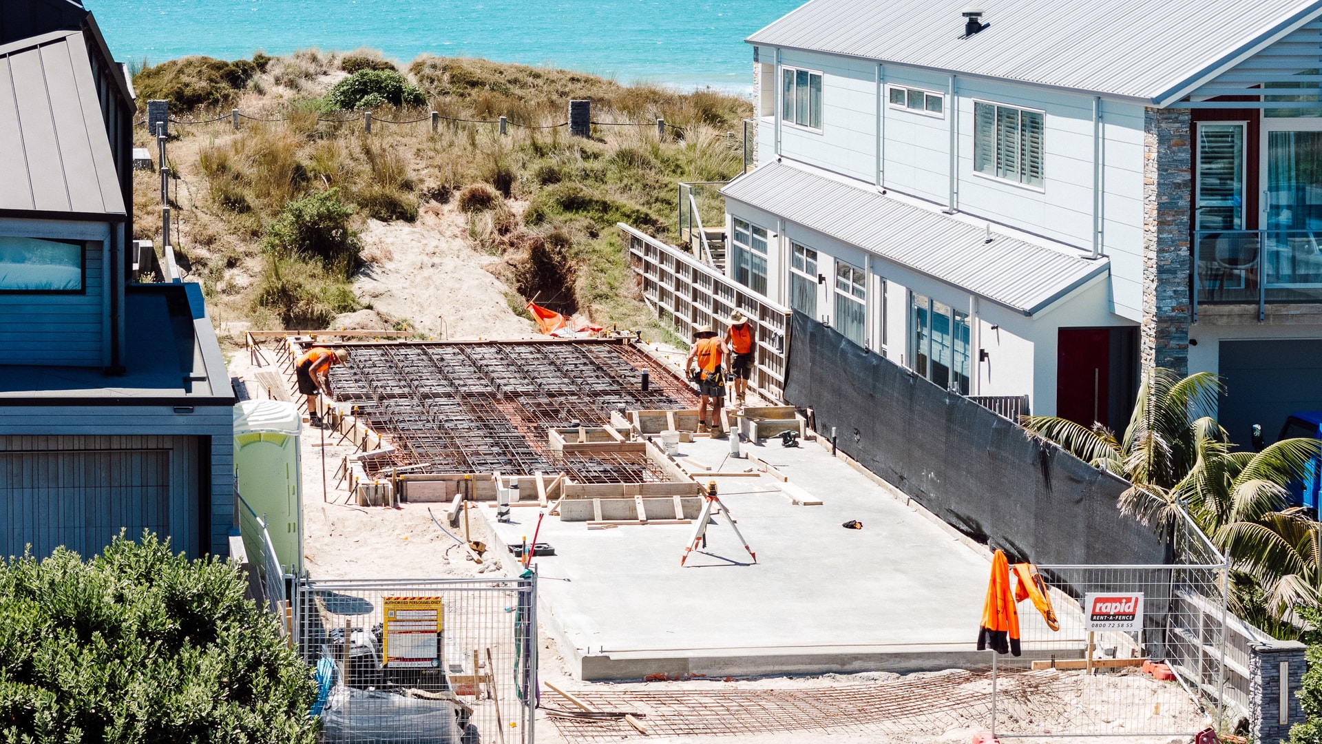 Construction site of a building foundation next to a modern house by the ocean, with workers in orange shirts and safety hats.
