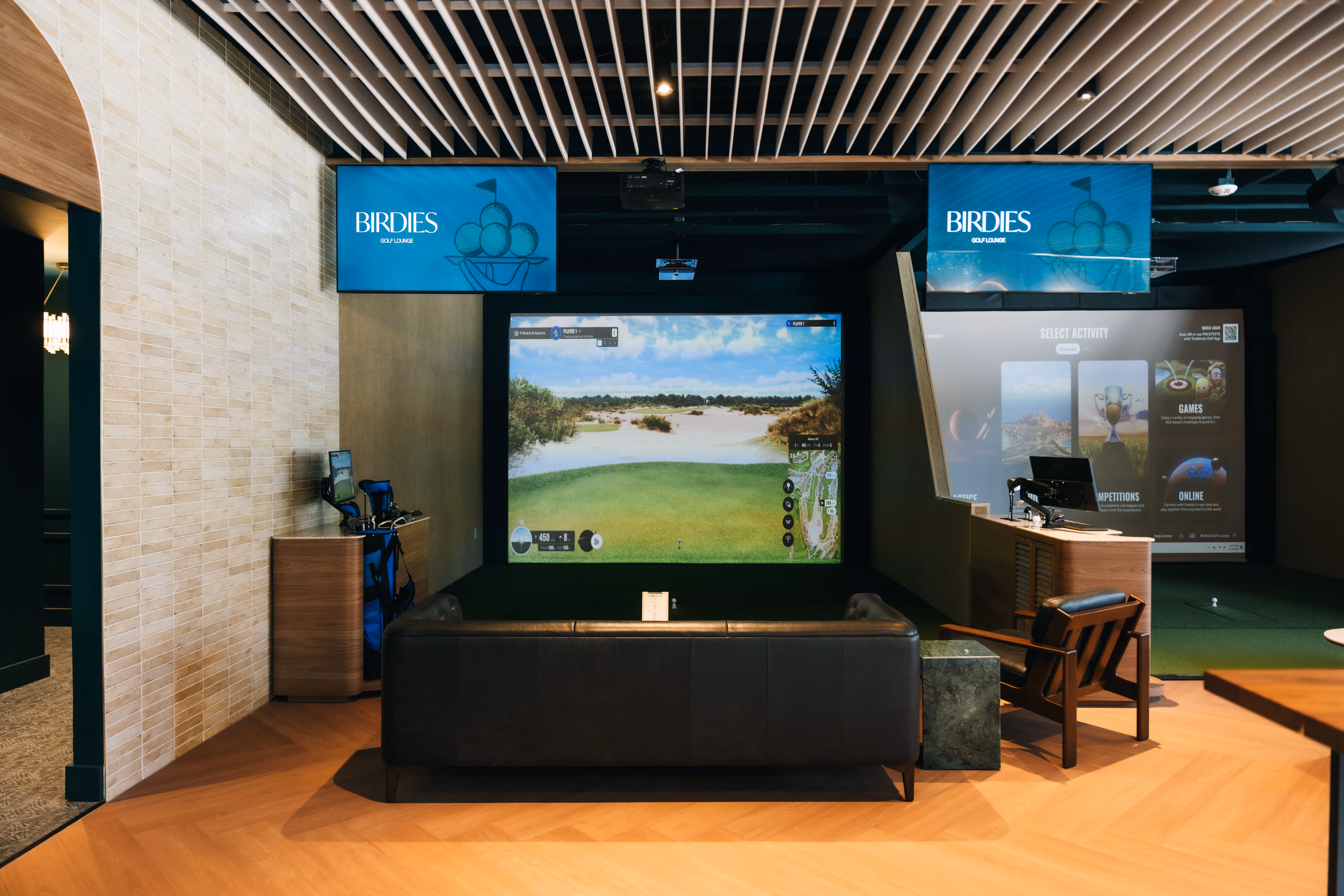 Indoor golf simulator lounge with a black leather sofa, wooden chair, and two screens displaying golf content.