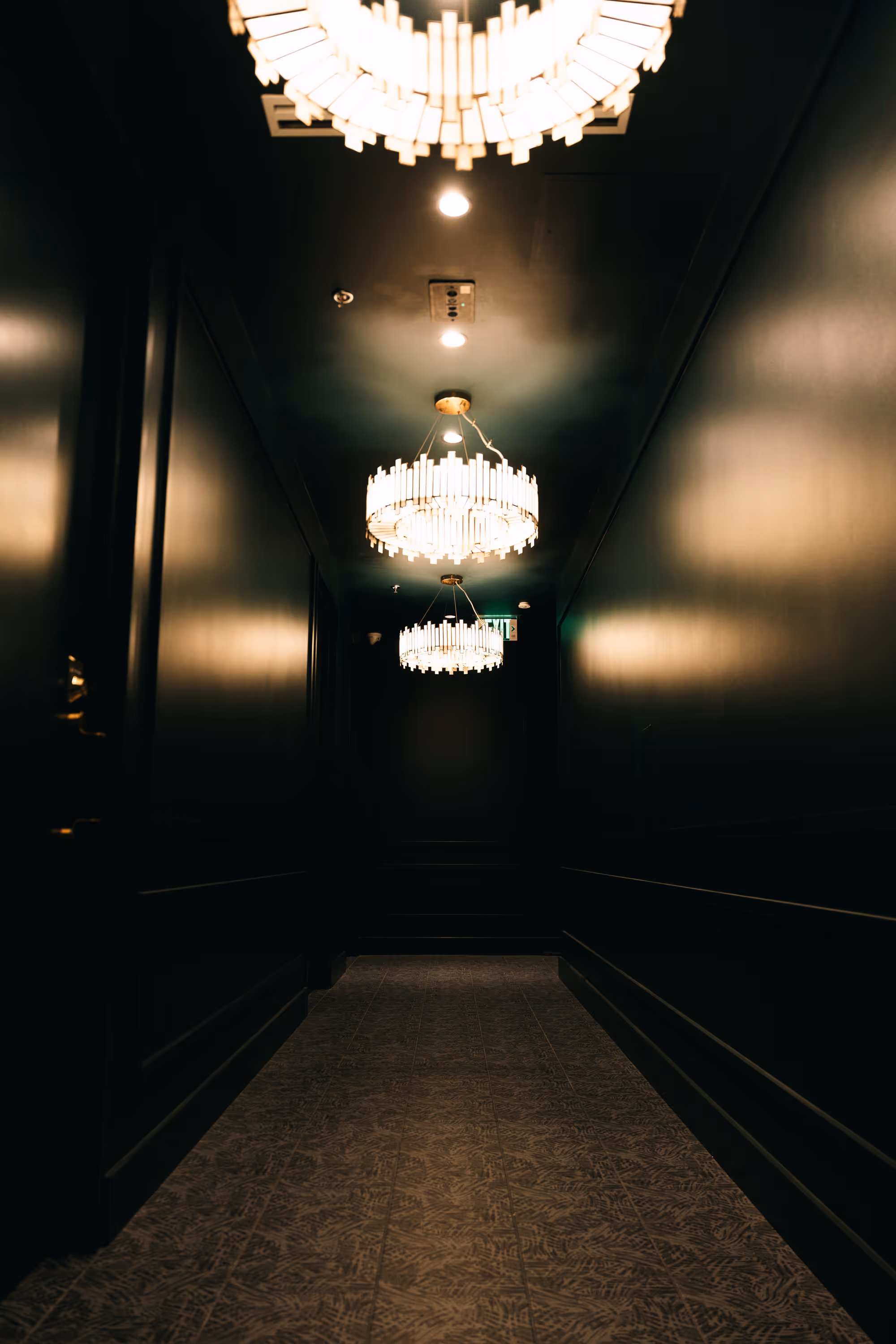 Dimly lit narrow hallway with ornate chandeliers hanging from the ceiling and patterned floor tiles.