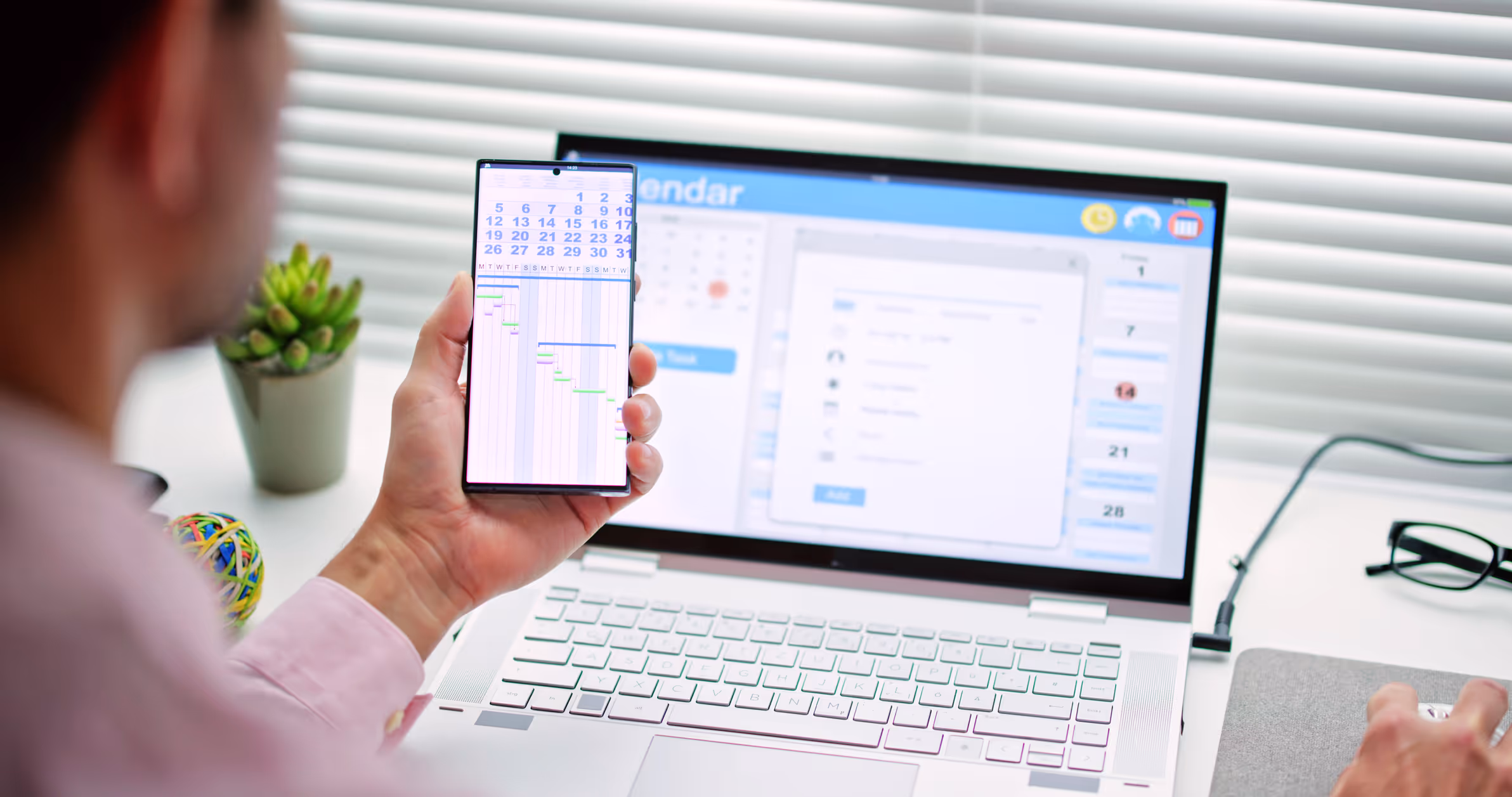 Person holding smartphone showing a calendar and project timeline in front of a laptop with a calendar app on the screen.