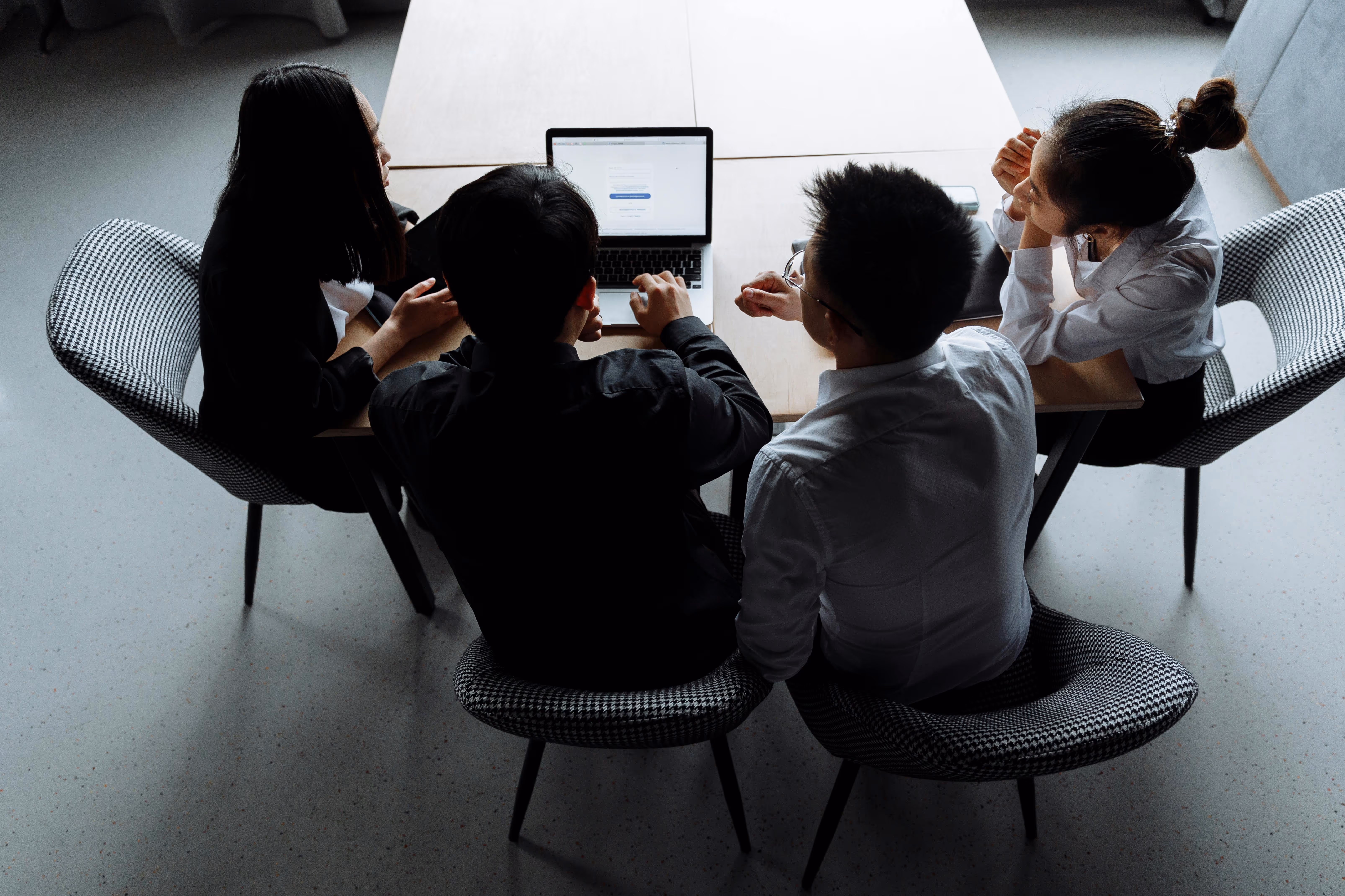 Four people seated around a table, viewing a laptop screen together in a modern office setting.
