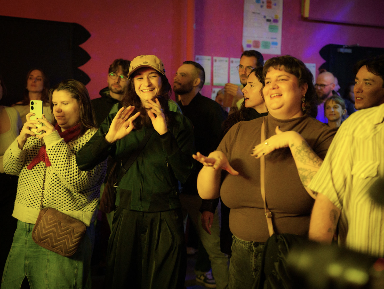 Groupe de personnes dans une salle avec éclairage jaune et rose, certaines souriant et une personne prenant une photo avec son téléphone.