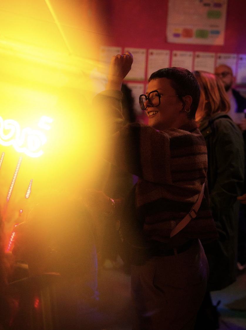Personne souriante avec lunettes dans une pièce sombre, illuminée par une lumière jaune intense à gauche.