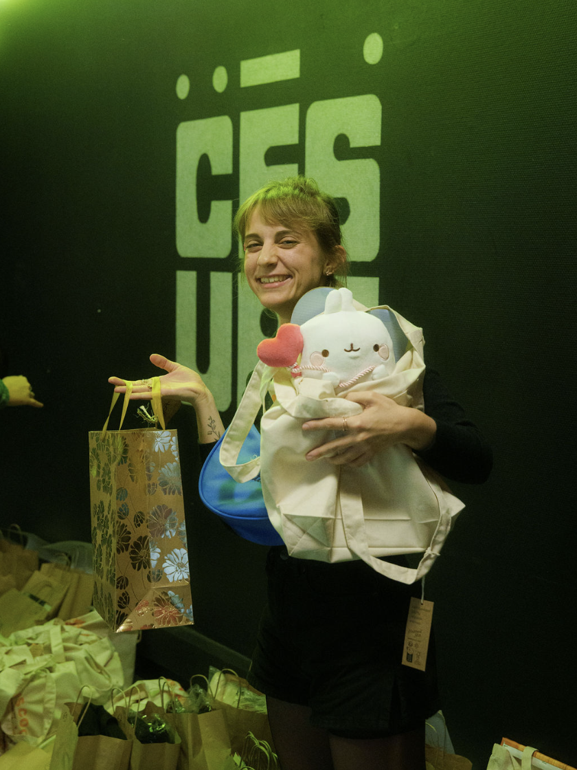Une femme souriante tenant un sac en papier à motifs floraux dorés et un sac en tissu blanc contenant une peluche blanche avec un cœur rose, devant un mur sombre avec un texte flou en arrière-plan.
