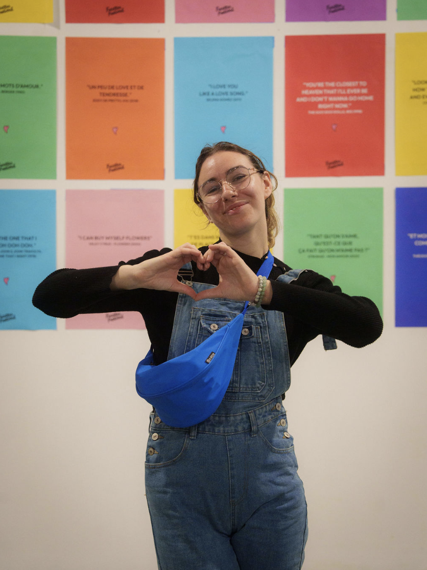 Jeune femme souriante en salopette en jean avec un sac bleu forme un cœur avec ses mains devant un mur décoré de posters colorés.