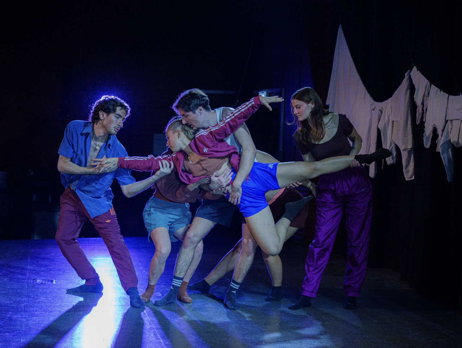 Quatre personnes en vêtements colorés exécutent une danse contemporaine en studio sombre avec un éclairage bleu, une suspendent les vêtements blancs sur une corde en arrière-plan.