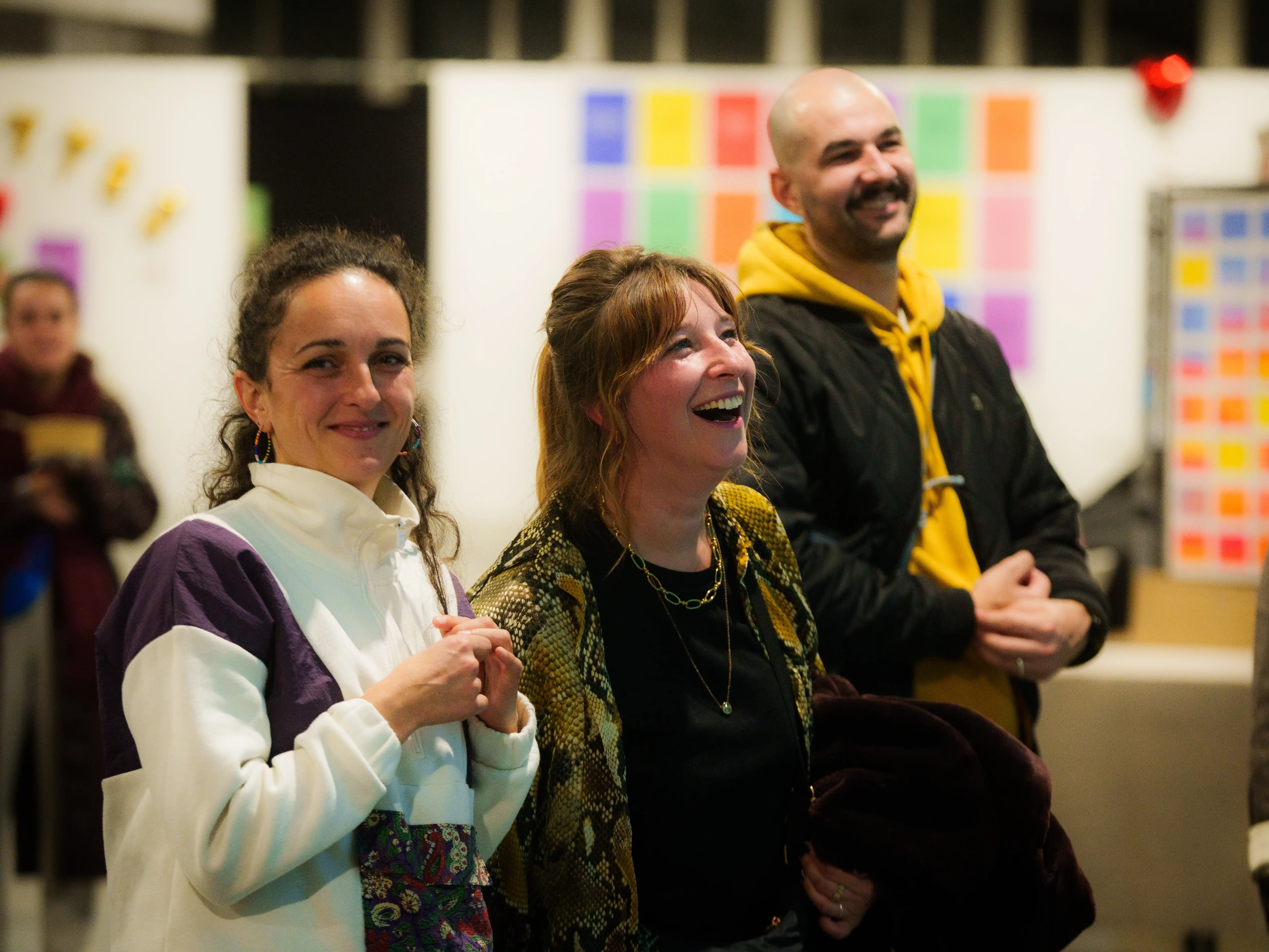 Trois adultes souriants regardant vers l'avant dans une pièce avec des tableaux colorés en arrière-plan.