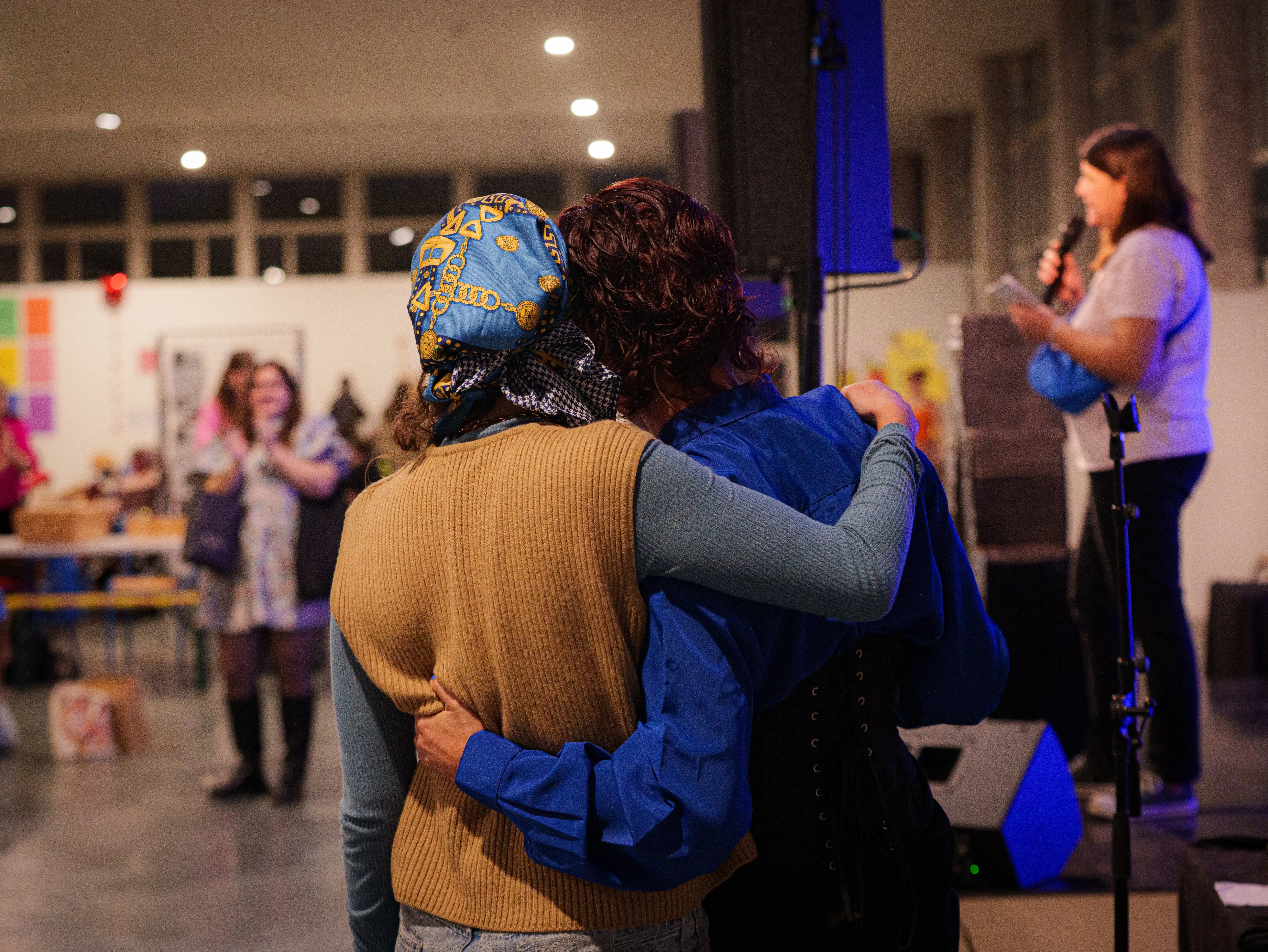 Deux personnes enlacées de dos lors d'un événement en intérieur avec une femme parlant au micro en arrière-plan.