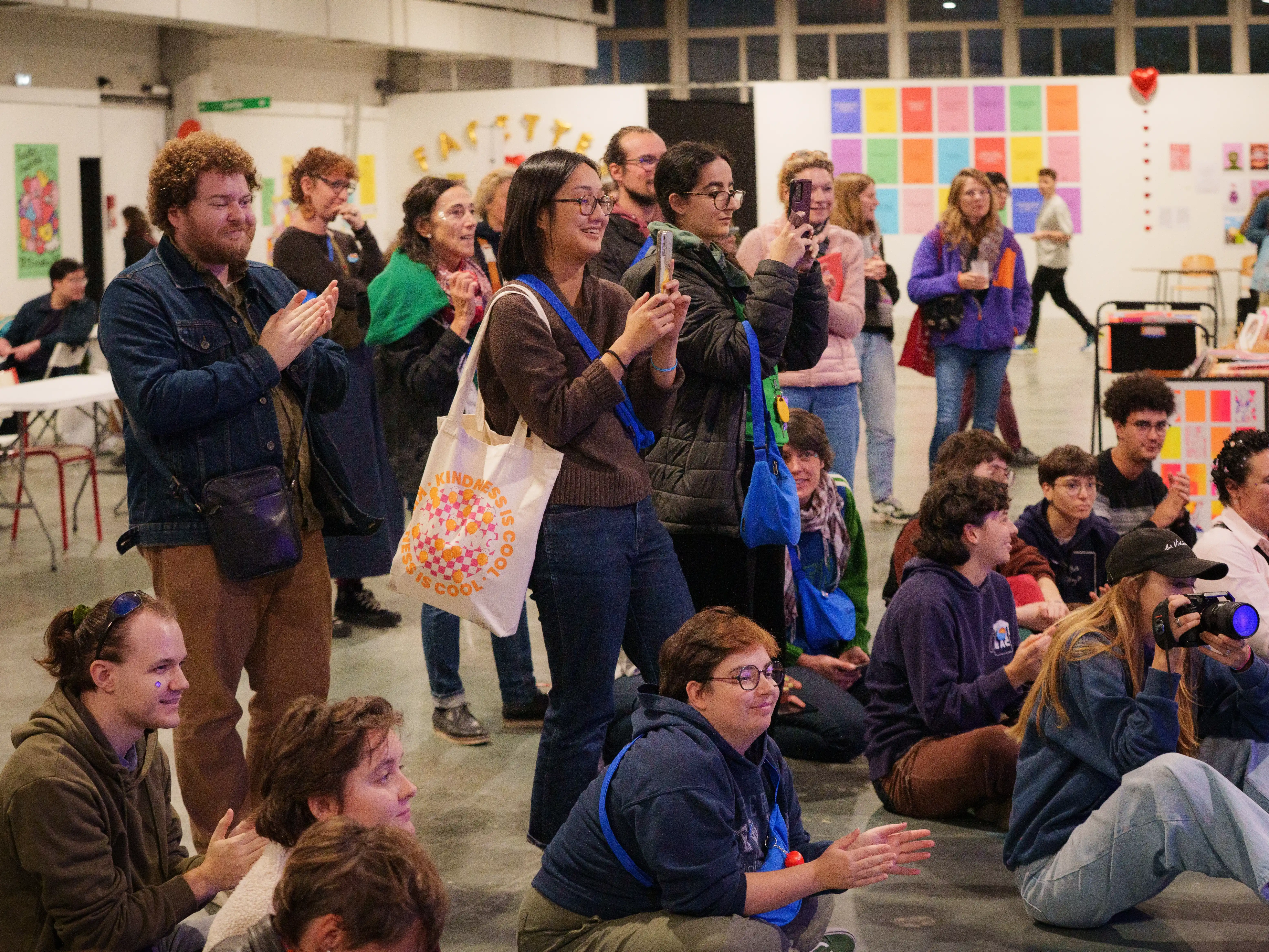 Groupe de personnes dans un espace intérieur, certaines debout et d'autres assises, applaudissant et prenant des photos avec leur téléphone.