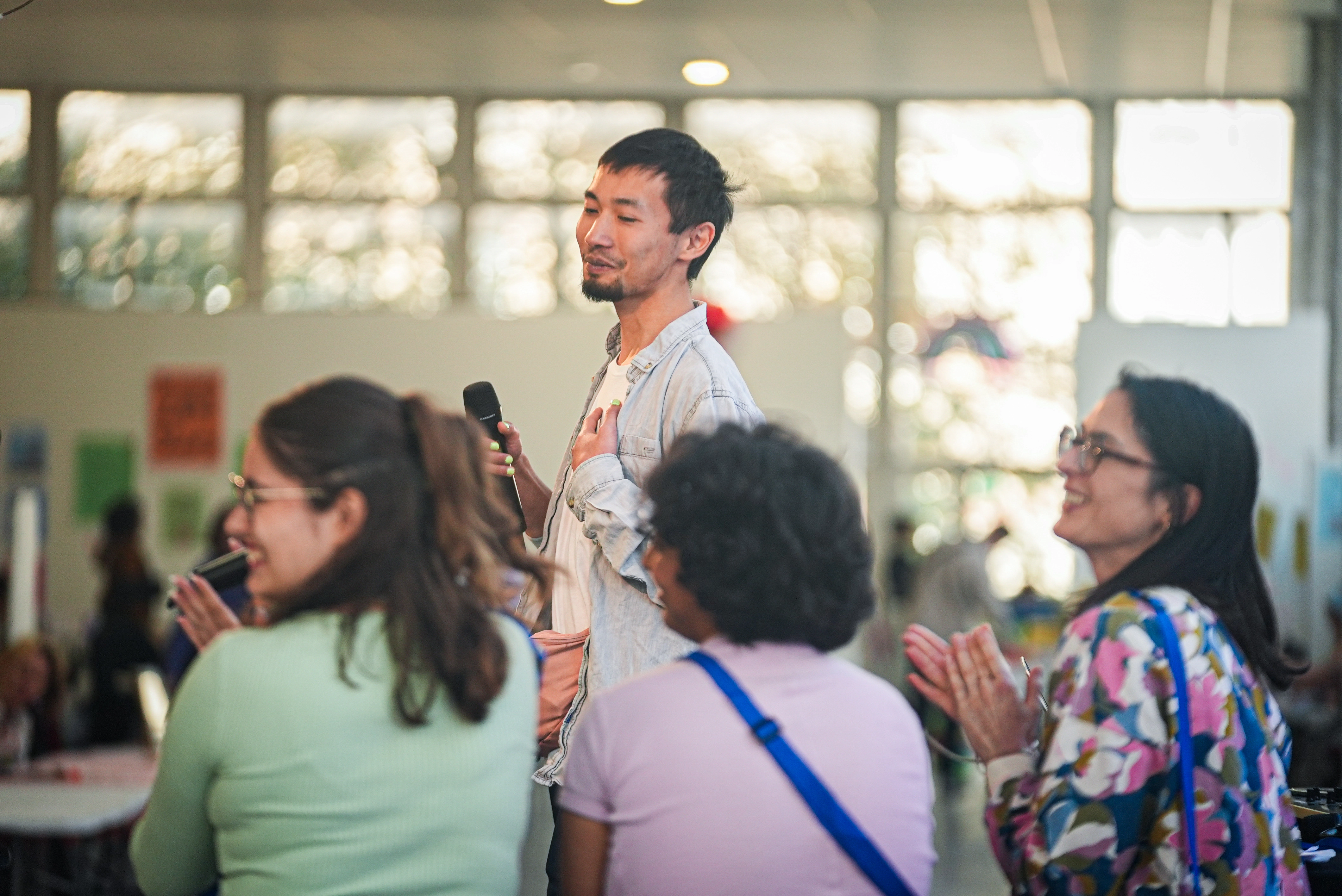 Un homme tenant un microphone parle devant un petit groupe de personnes assises qui l'applaudissent dans une salle lumineuse.