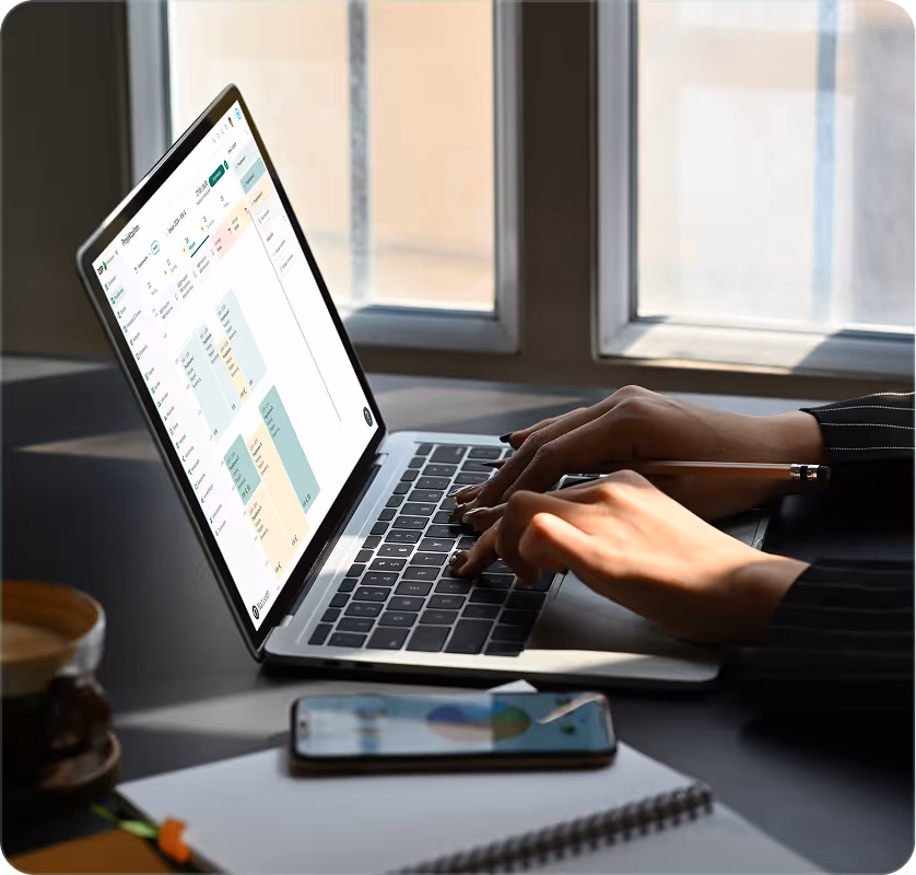 Person tippt auf einem Laptop mit Finanz- oder Budgetplanungs-Software neben einem Smartphone und Notizbuch auf einem Tisch.