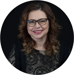 Un portret al unei femei zâmbitoare cu păr ondulat și ochelari, îmbrăcată într-o bluză cu model negru, cu un fundal întunecat, oferind o imagine profesională și caldă.




