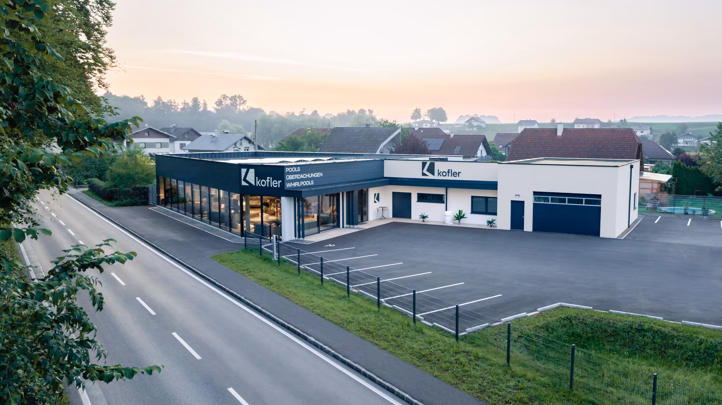 Geschäftsgebäude von Kofler mit großen Glasfenstern und leerem Parkplatz neben einer zweispurigen Straße.