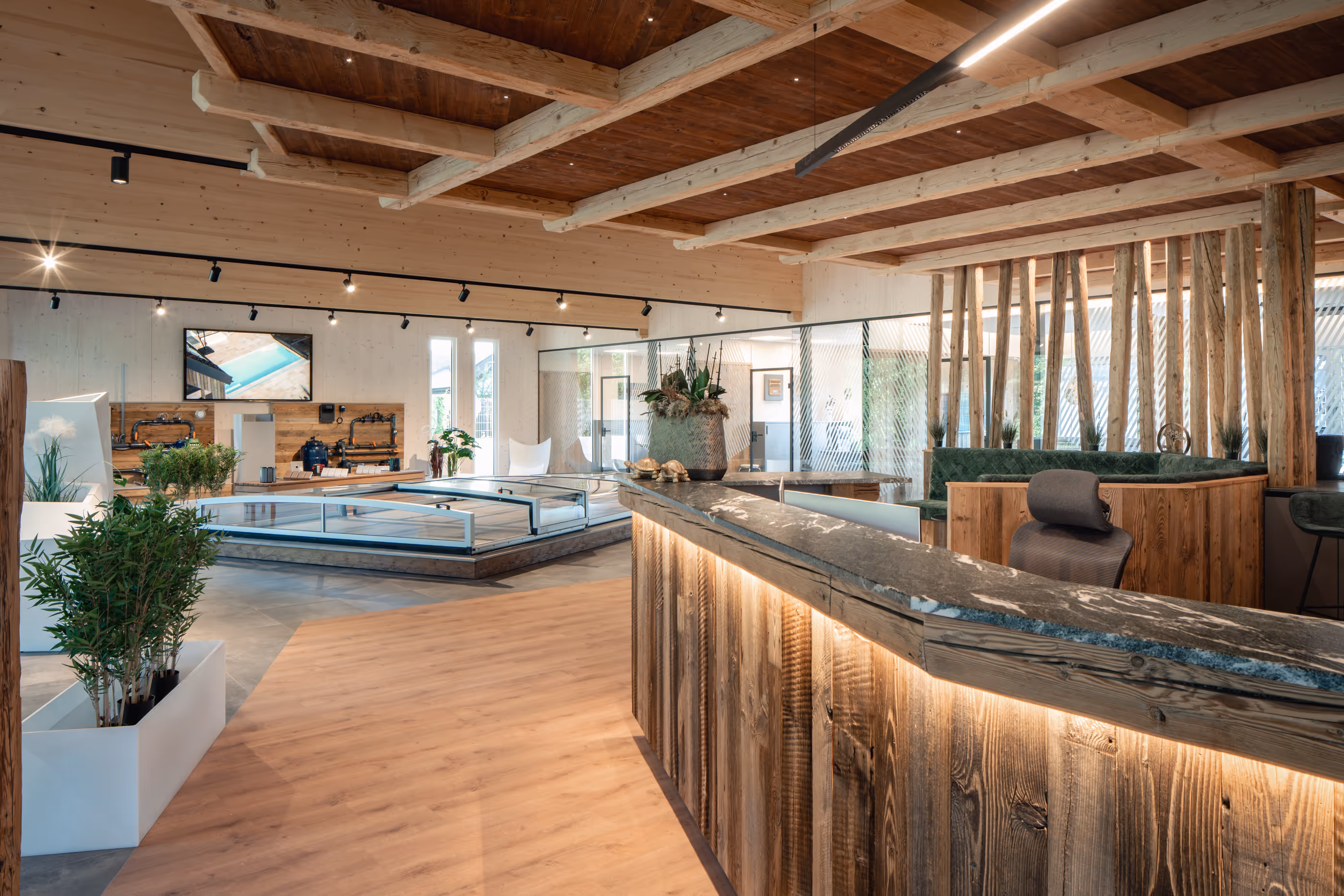 Moderner Schauraum mit Holzbalkendecke, beigem Holzboden, einem beleuchteten Empfangstresen aus Holz, Pflanzen und Sitzbereichen mit grünen Polstern.