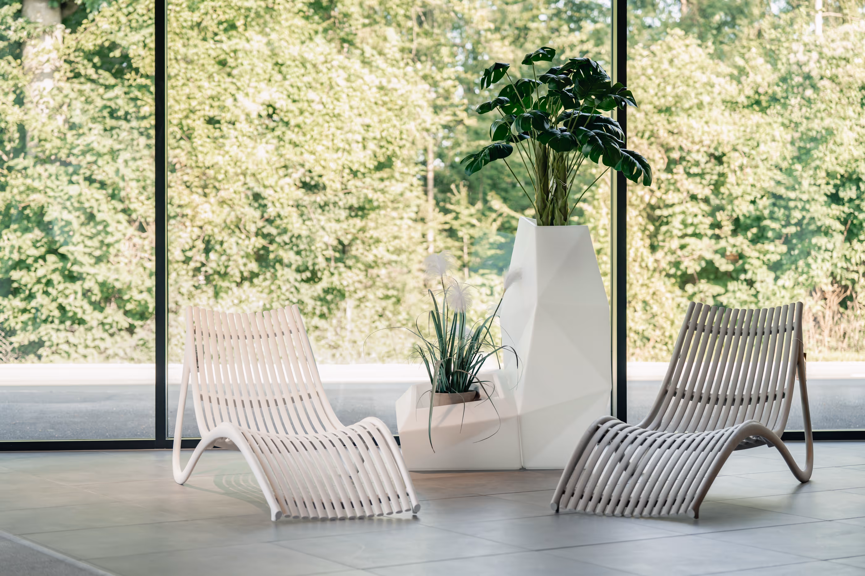 Zwei weiße moderne Liegestühle aus gebogenen Holzleisten vor großen Fenstern mit Blick auf grünes Laub, daneben zwei weiße geometrische Pflanzentöpfe mit grünen Pflanzen.