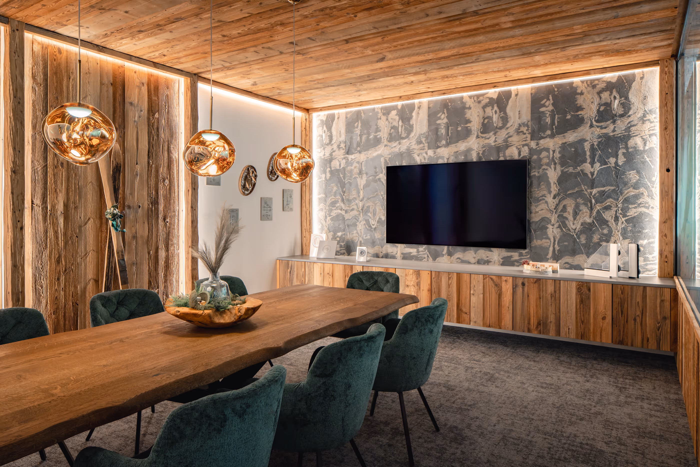Moderner Besprechungsraum mit großem Holztisch, grünen Samtstühlen, drei kupferfarbenen Hängependelleuchten und großem Fernseher an einer Wand mit Textur.