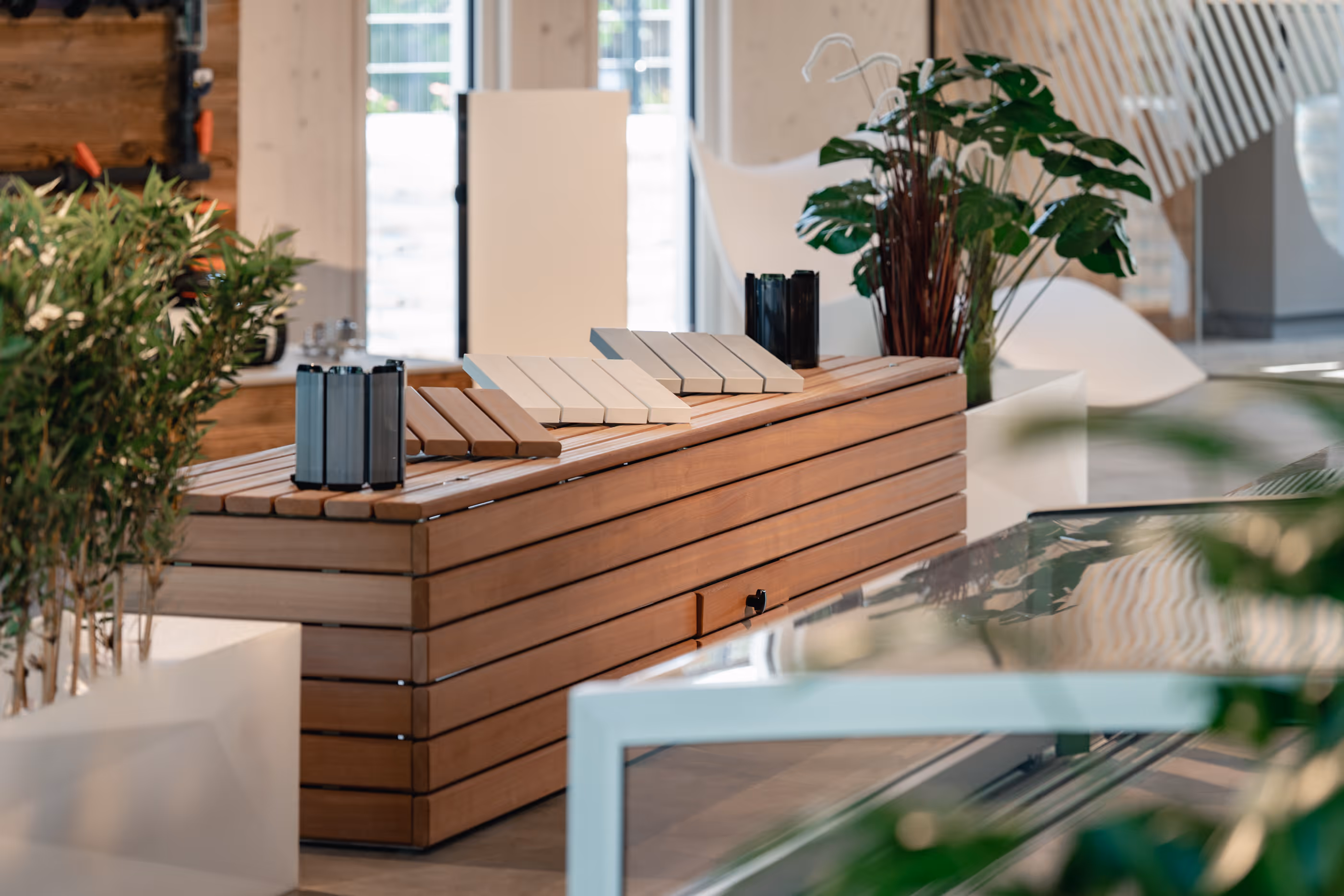 Moderner Innenraum mit einem langen Holztisch, auf dem verschiedene Farbproben liegen, umgeben von grünen Pflanzen und weißen Möbeln.