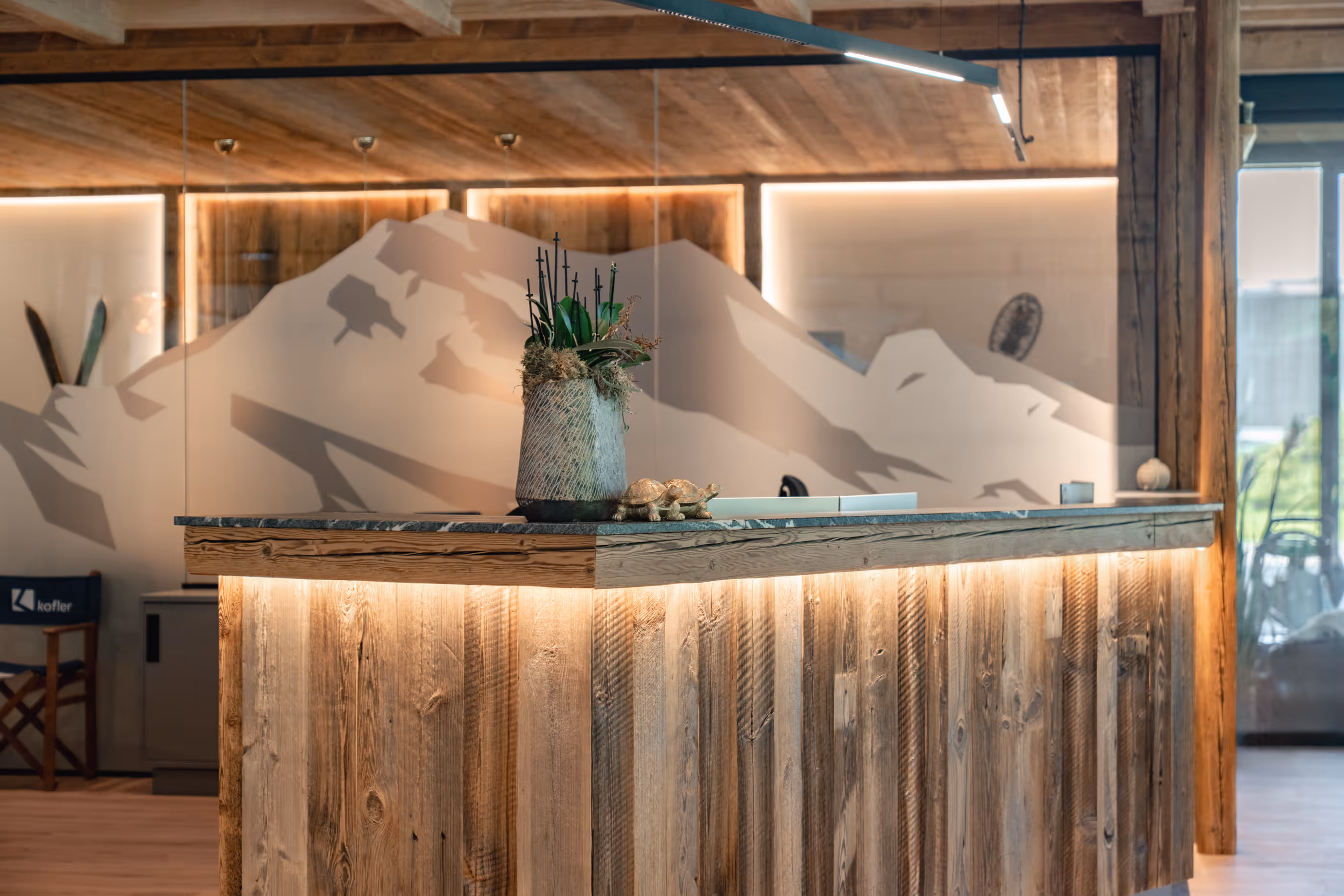 Rustikaler Empfangstresen aus Holz mit Pflanzenarrangement und Schreibtisch im Hintergrund vor einer Glaswand mit Berggrafik.