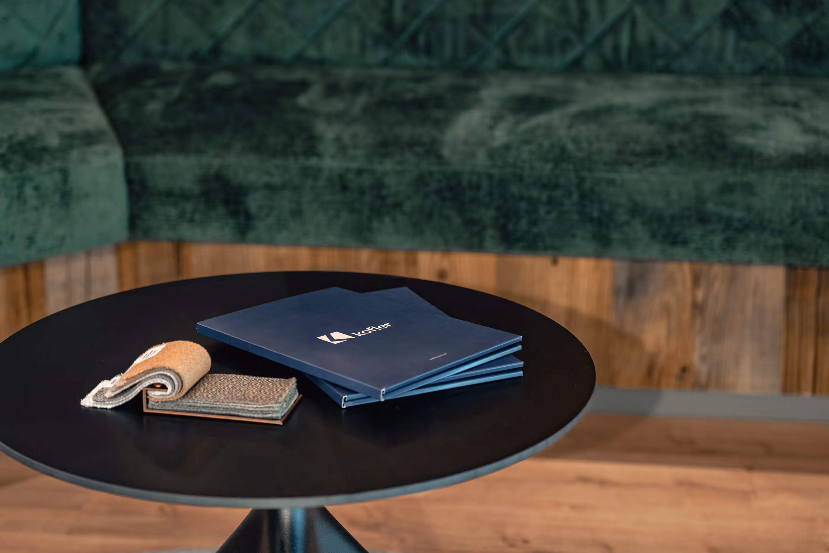Runder schwarzer Tisch mit drei blauen Ordnern mit dem Logo Kofler und zwei Musteretiketten vor einem grünen Samtsofa vor Holzwand und Holzboden.