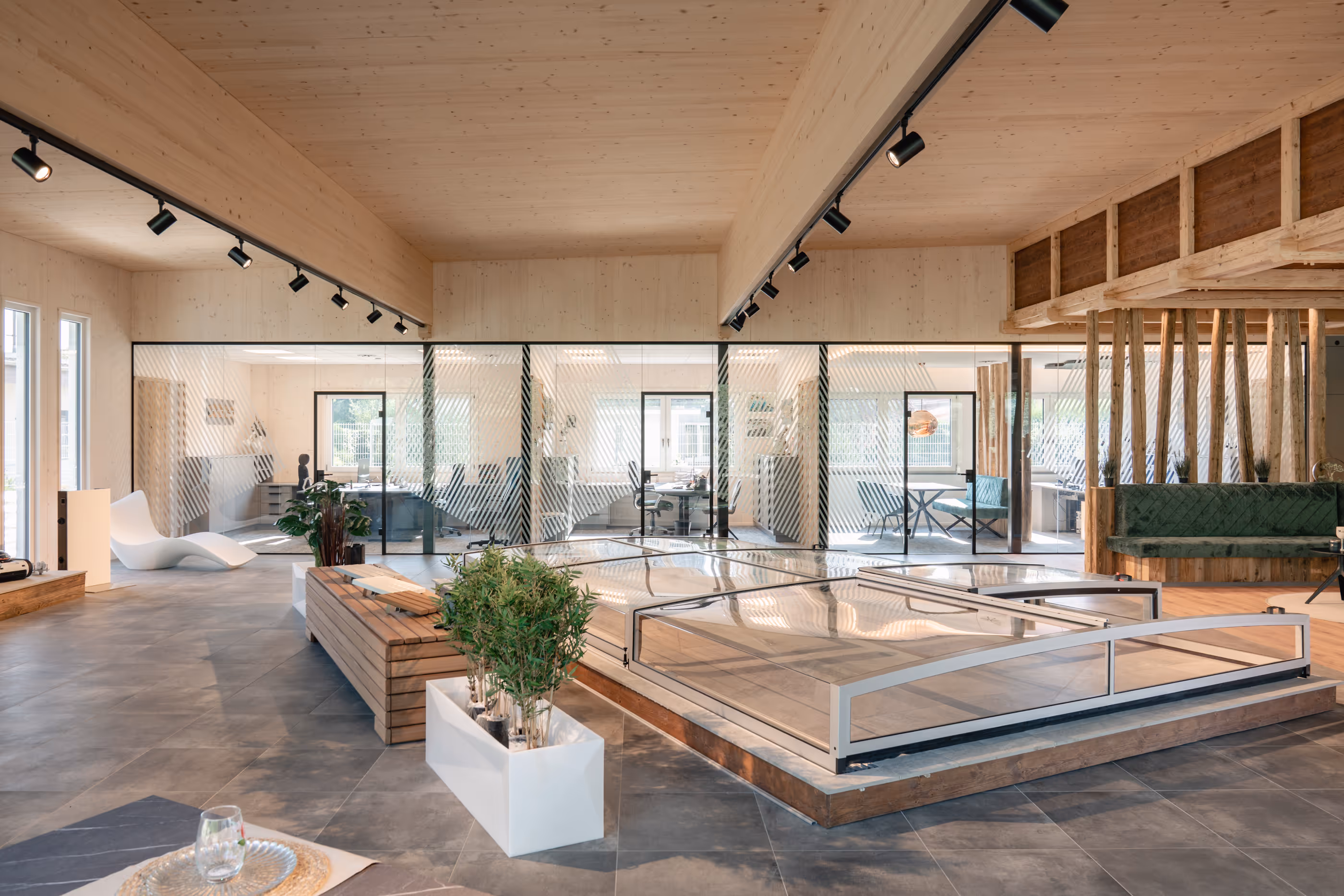 Moderner, offener Büroraum mit Glaswänden, Holzbalkendecke, Pflanzen und Sitzecke mit grüner Polsterbank.