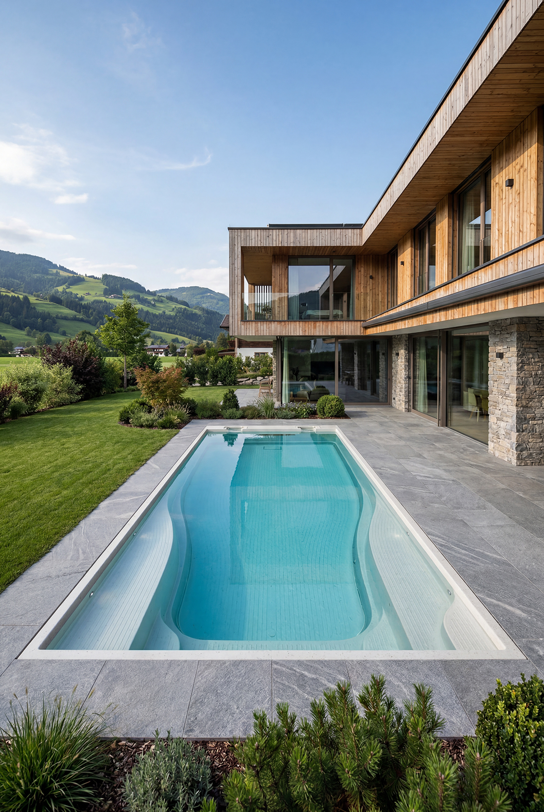Moderner Holzhaus mit Terrasse, Pool und grüner Landschaft vor bewaldeten Hügeln unter blauem Himmel.
