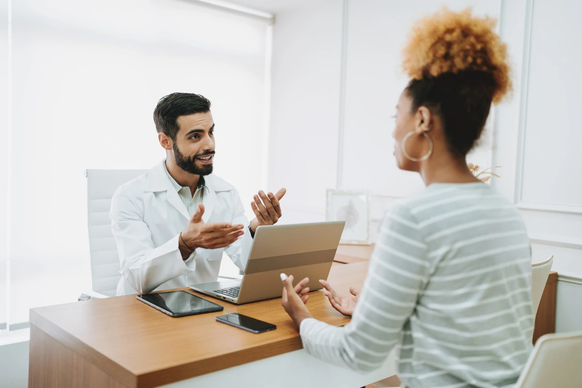 Ein Arzt sitzt an einem Schreibtisch und spricht freundlich mit einer Patientin. Vor ihm stehen ein Laptop, ein Tablet und ein Smartphone. Beide gestikulieren während des Gesprächs und wirken konzentriert und engagiert.
