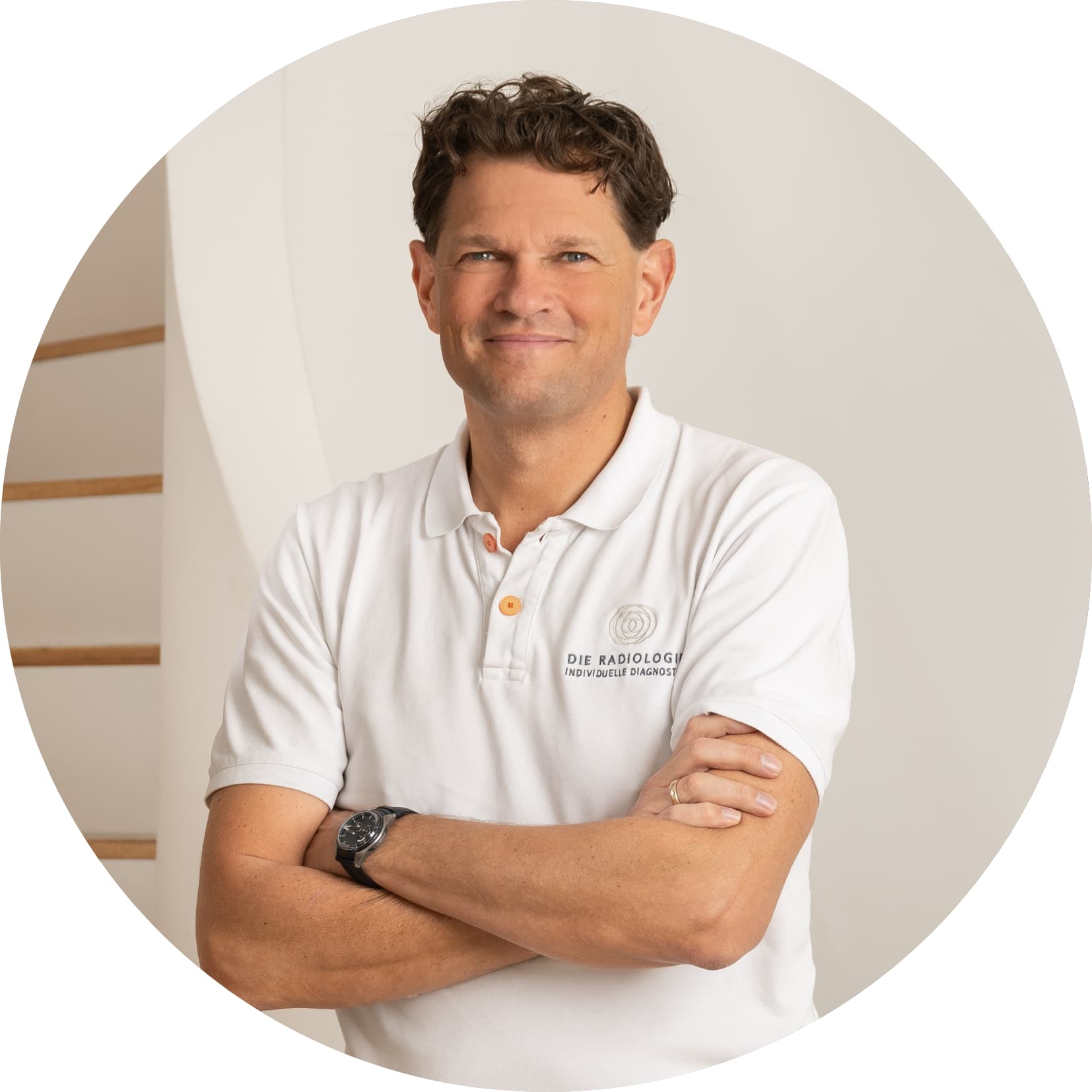 Porträt eines lächelnden Mannes in weißem Polohemd mit verschränkten Armen, vor einem hellen, minimalistischen Hintergrund.