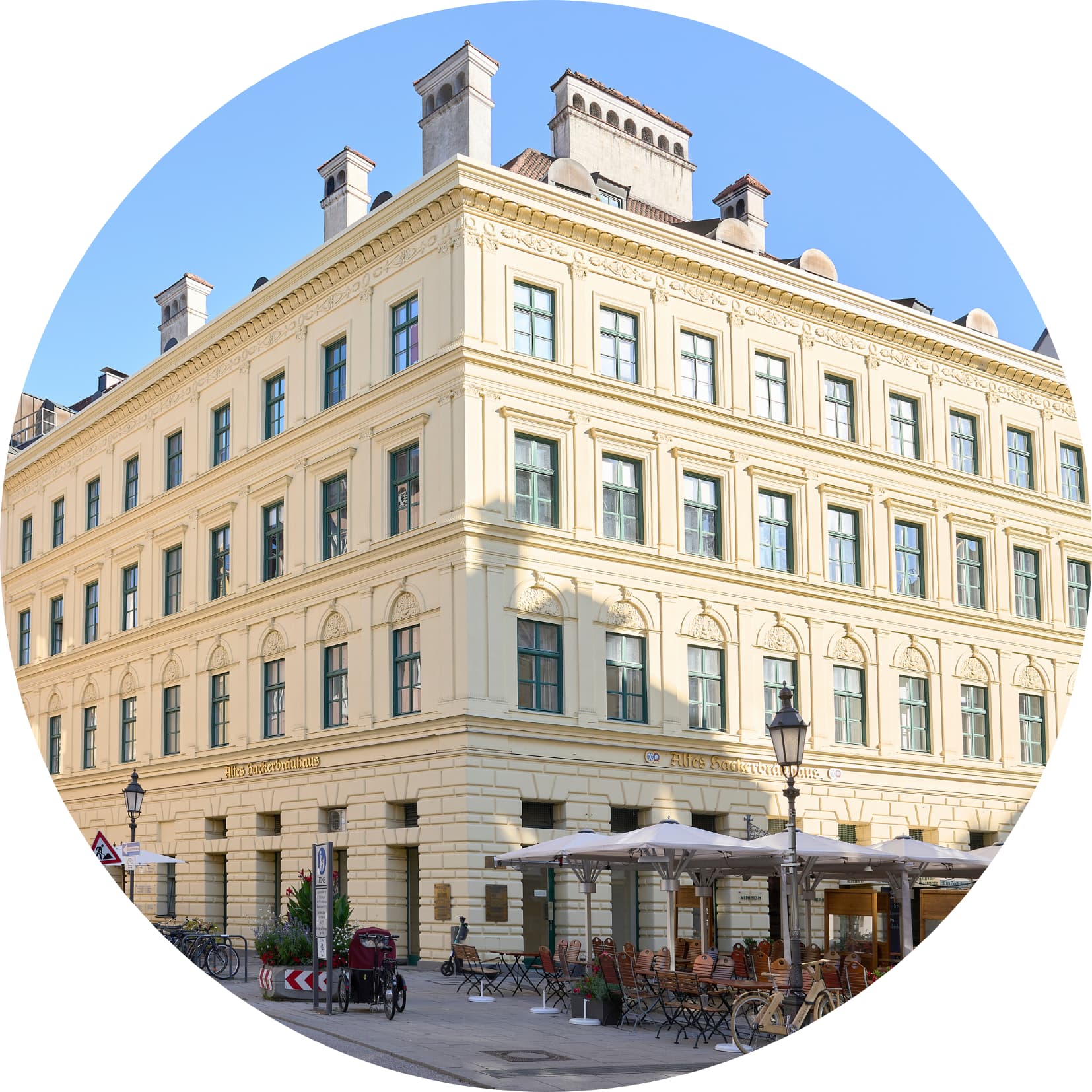 Historisches, hellgelbes Eckgebäude mit mehreren Stockwerken, grünen Fensterrahmen und verzierten Fassadendetails; davor ein Außenbereich mit Tischen, Stühlen und Sonnenschirmen an einer ruhigen Straßenkreuzung.