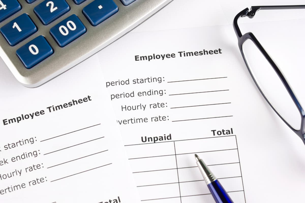 Employee timesheet forms lying on a desk next to a calculator, a pair of glasses, and a pen.