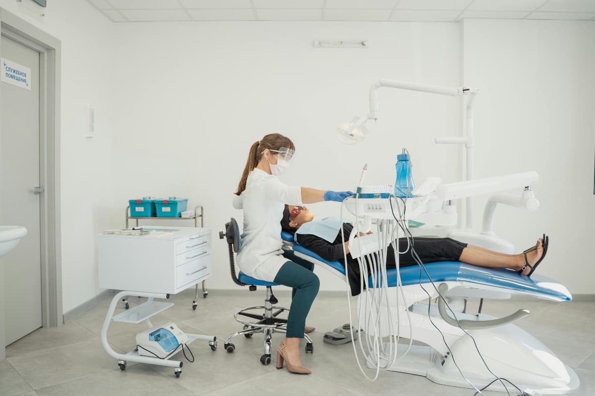 Eine Zahnärztin mit Schutzmaske und Gesichtsschild behandelt eine liegende Patientin in einem modernen, hellen Behandlungsraum. Um sie herum stehen zahnmedizinische Geräte, Instrumente und Schläuche; im Hintergrund sind Schränke und medizinische Behälter.