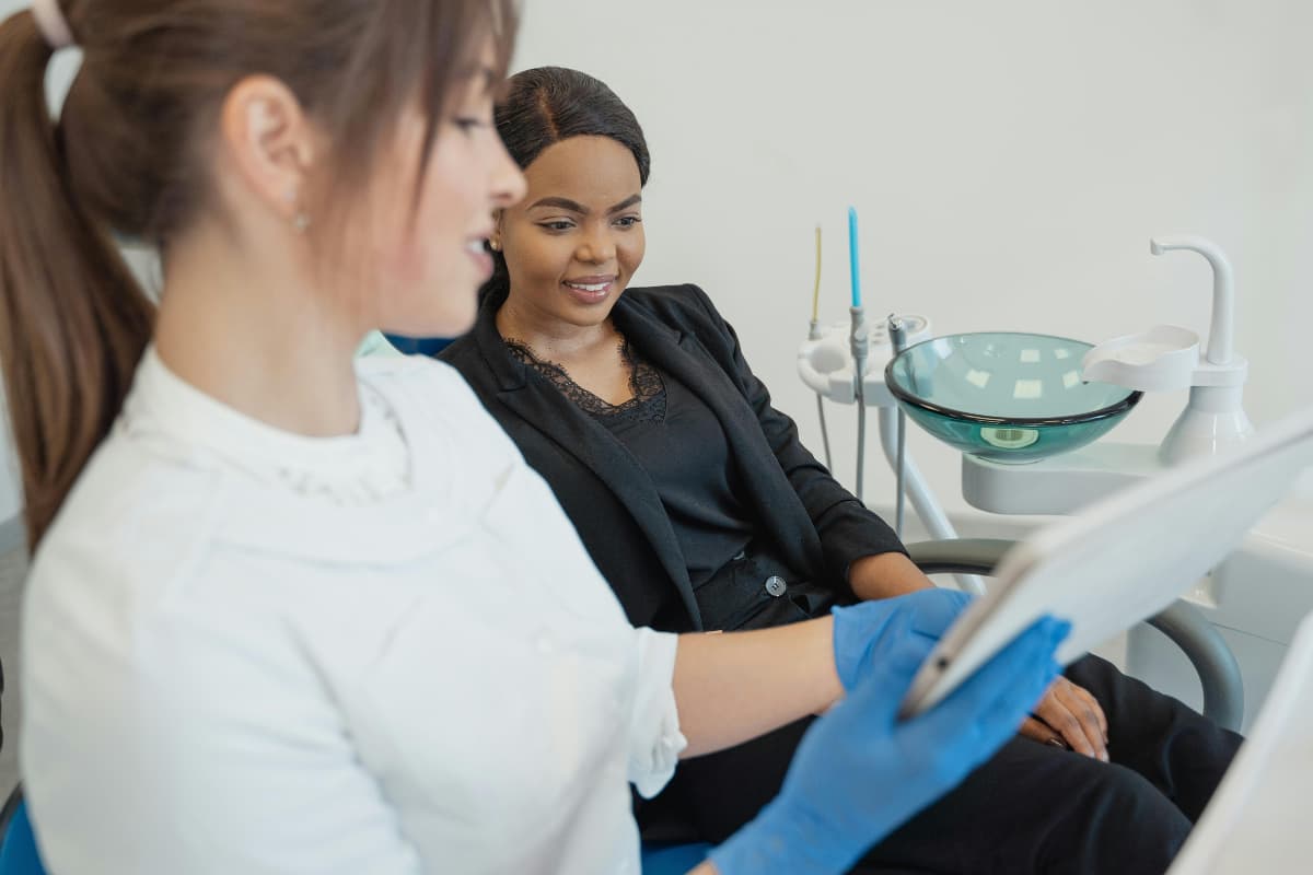 Eine Zahnärztin zeigt einer Patientin auf einem Tablet Informationen, während beide in einem modernen Behandlungszimmer sitzen. Die Patientin lächelt interessiert, im Hintergrund sind zahnmedizinische Geräte und ein Glaswaschbecken zu sehen.