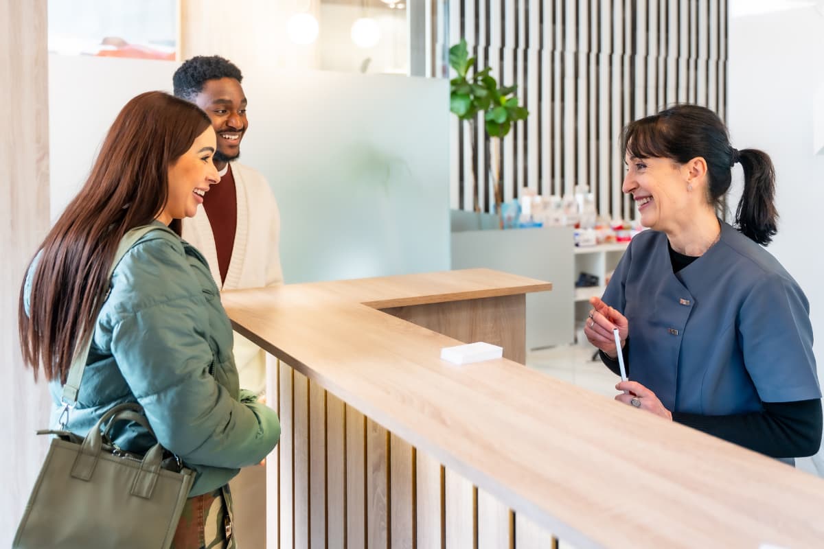 Eine medizinische Fachangestellte steht lächelnd hinter dem Empfangstresen einer Praxis und begrüßt ein lachendes Paar, das gerade ankommt. Alle wirken freundlich und entspannt in der modernen, hellen Praxiseingangssituation.