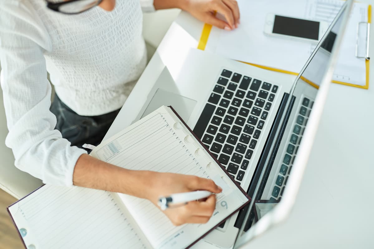 Eine Person sitzt an einem Schreibtisch, schreibt in einen geöffneten Kalender und arbeitet gleichzeitig an einem Laptop. Auf dem Tisch liegen ein Smartphone und Unterlagen. Die Szene wirkt organisiert und konzentriert.