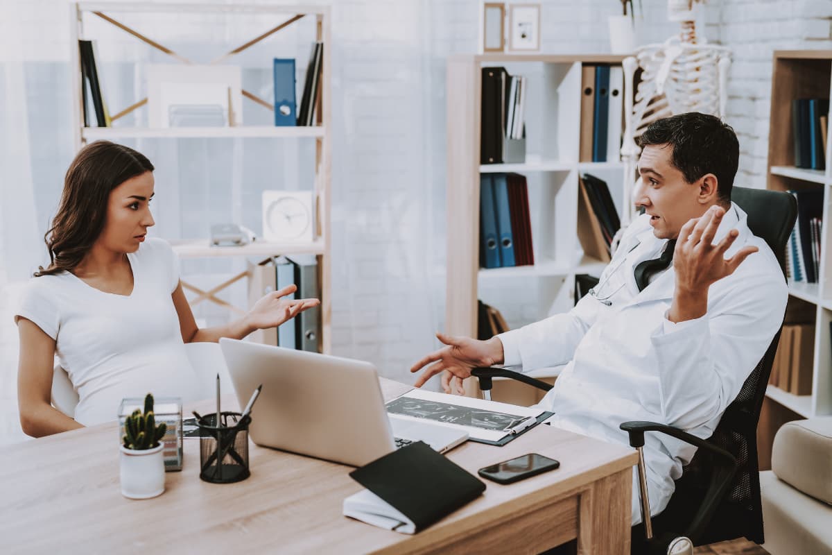 Ärztin und Arzt sitzen sich in einem Büro gegenüber und führen ein Beratungsgespräch an einem Schreibtisch mit Laptop und Unterlagen.