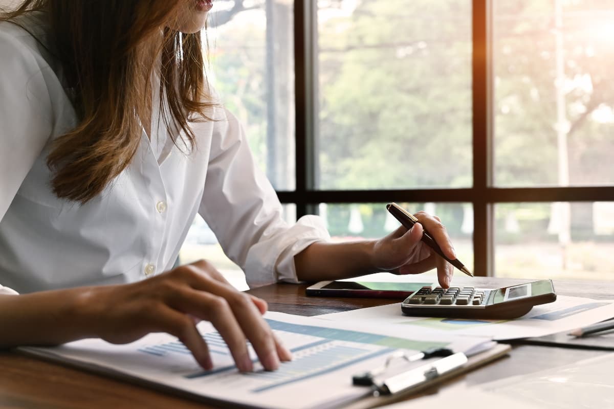 Person sitzt an einem Tisch und berechnet Finanzdaten mit einem Taschenrechner, während sie Dokumente und Diagramme prüft.