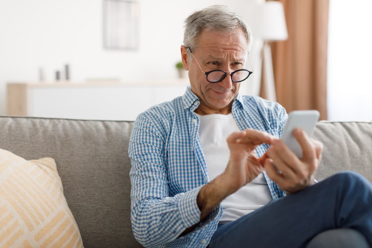 Older patient sitting at home on a sofa, using a smartphone to read or complete digital information.