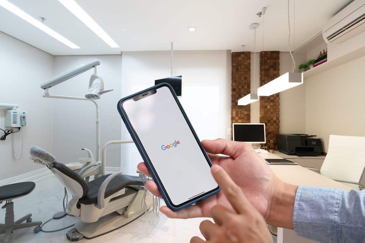 Hand hält ein Smartphone mit Google-Startseite in einer modernen Zahnarztpraxis mit Behandlungsstuhl und Geräten im Hintergrund.