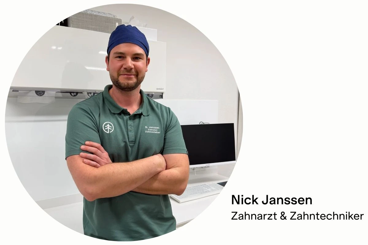 Porträt von Nick Janssen, Zahnarzt und Zahntechniker, in einer modernen Zahnarztpraxis. Er steht mit verschränkten Armen vor einem Behandlungsraum und trägt grüne Praxiskleidung sowie eine blaue OP-Haube.