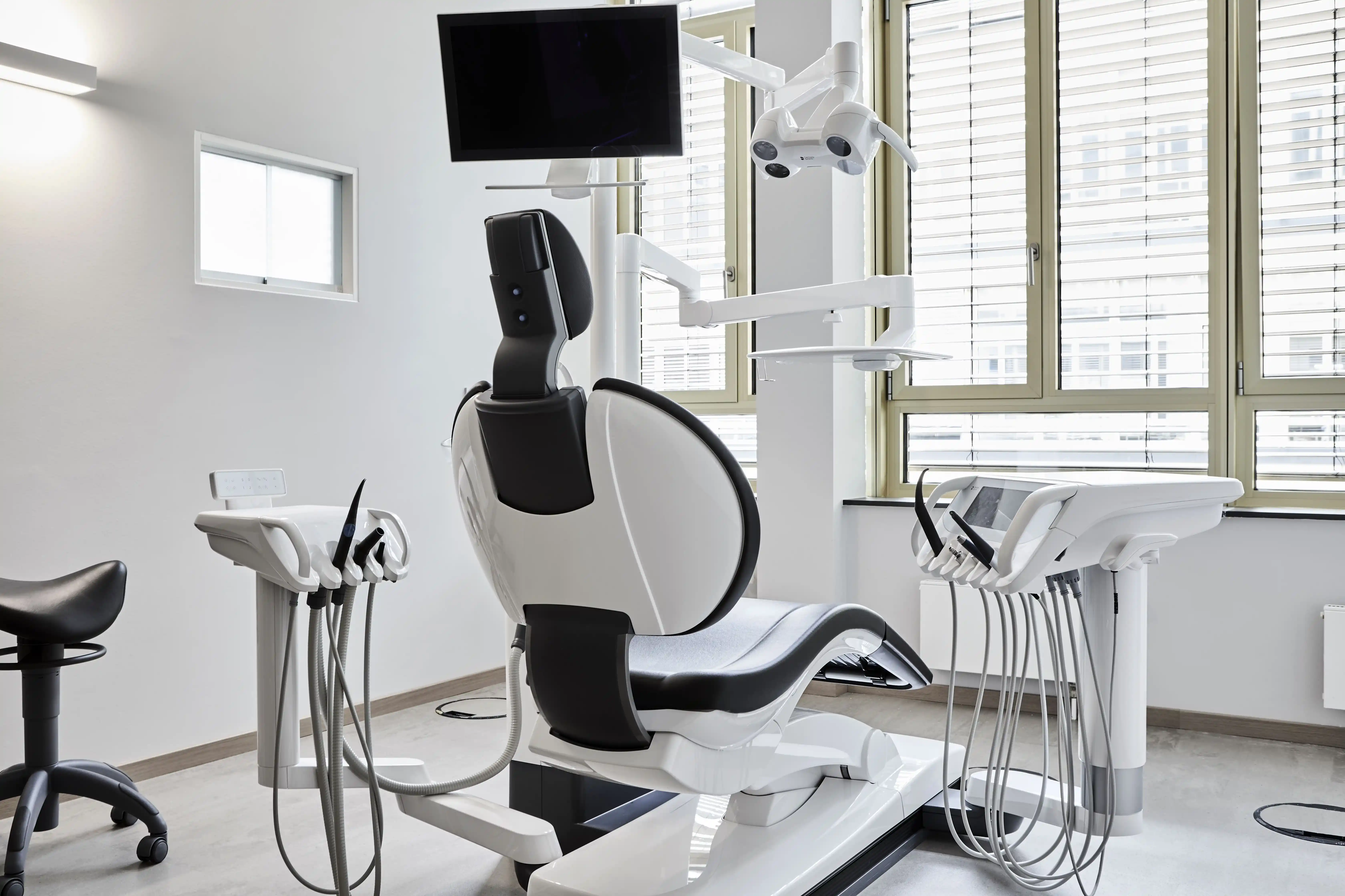 Modernes Behandlungszimmer in einer Zahnarztpraxis mit Zahnarztstuhl, Instrumenteneinheit, Monitor und großer Fensterfront.