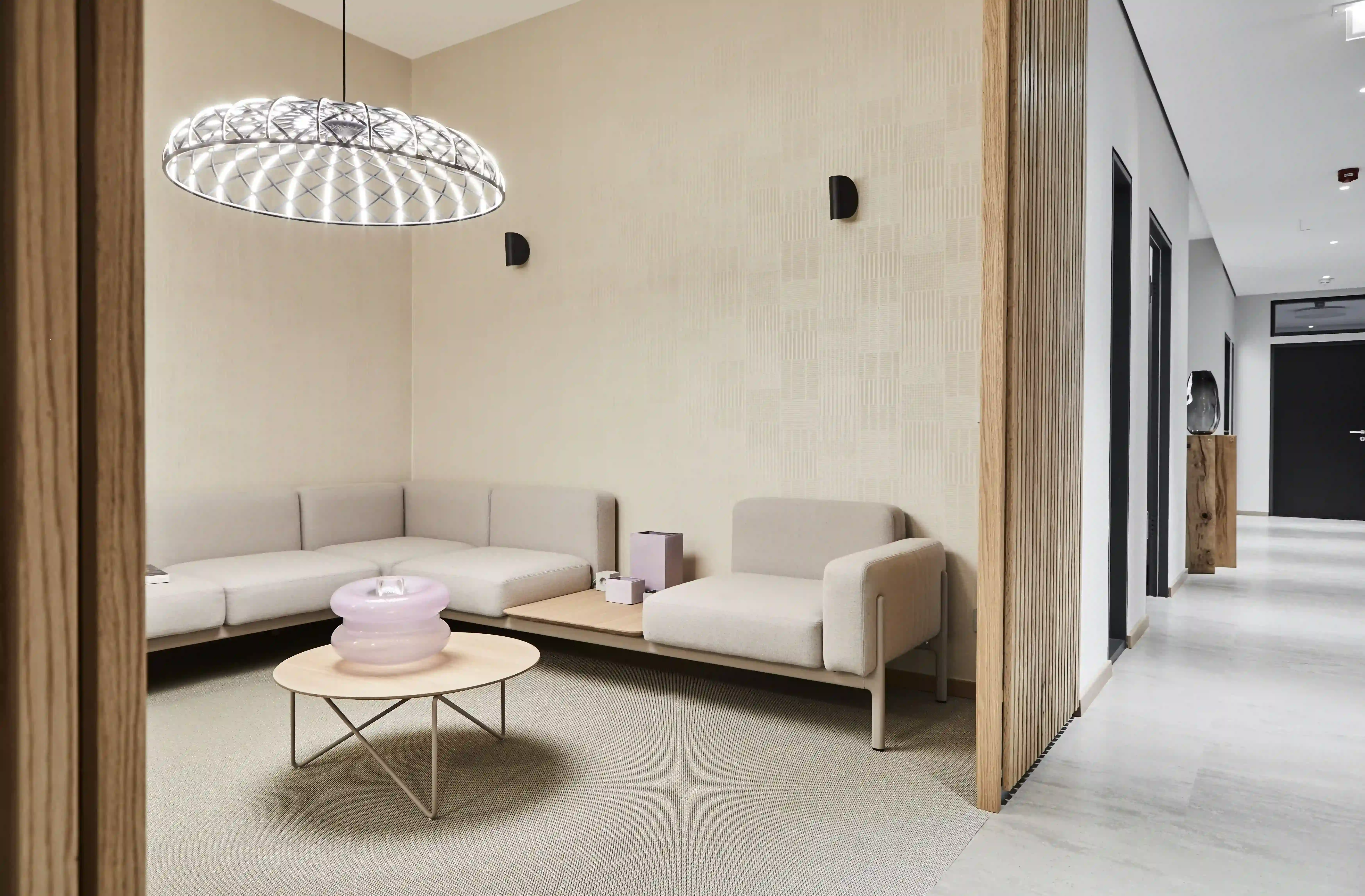 Moderner Wartebereich einer Zahnarztpraxis mit hellen Sitzmöbeln, rundem Beistelltisch und großer dekorativer Deckenleuchte in warmen Beige- und Holztönen.