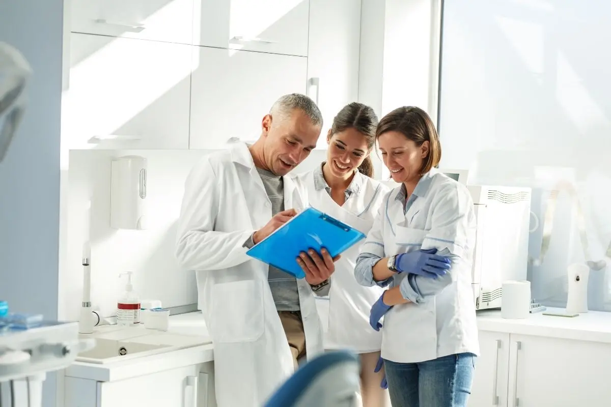 Three dental professionals in white lab coats standing together in a bright dental practice, smiling while reviewing documents on a blue clipboard. One person is wearing blue latex gloves.